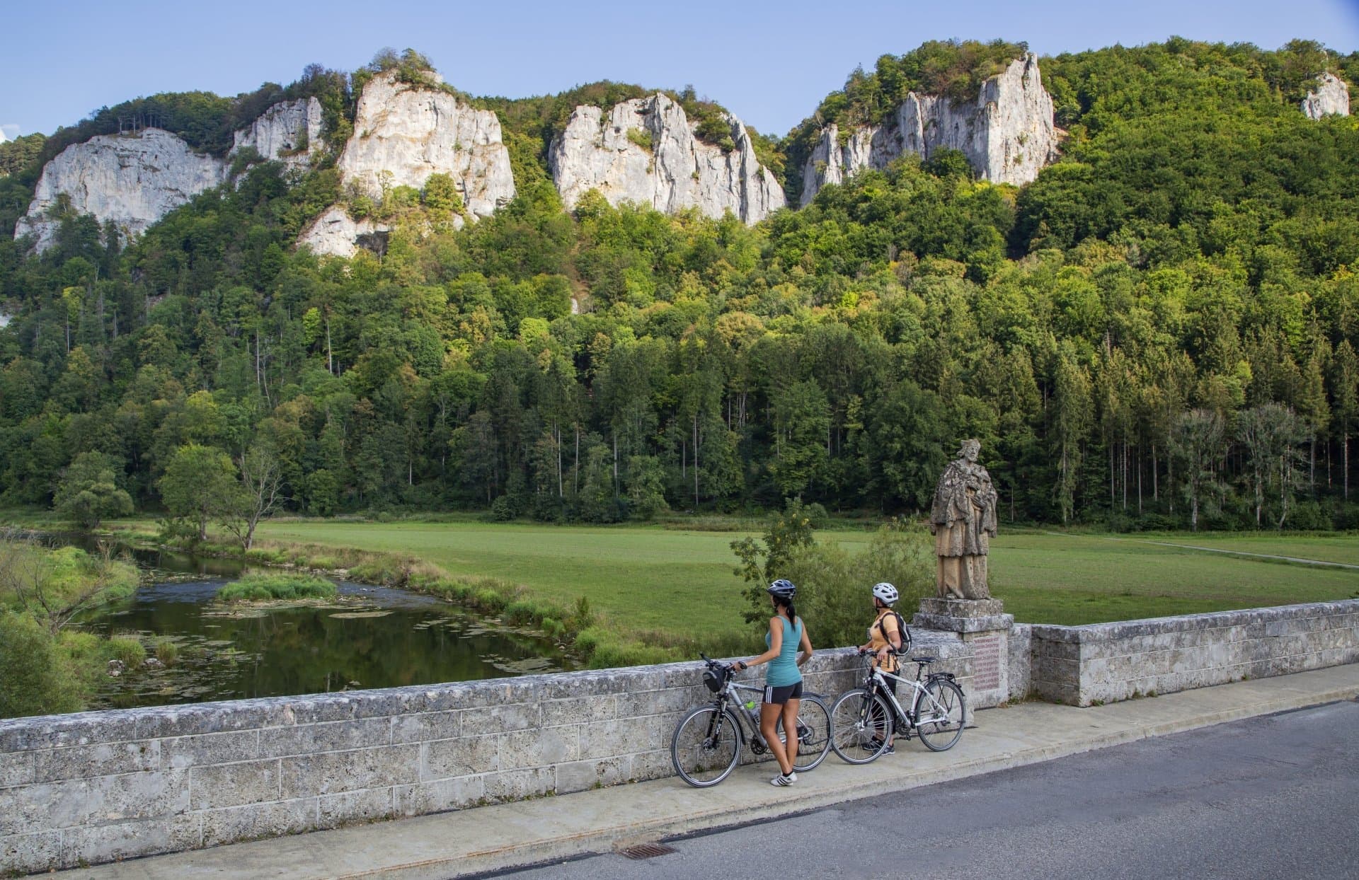 Zwei Radfahrerinnen auf einer Steinbrücke über die Donau neben einer Statue des Brückenheiligen Nepomuk  schauen in das von Felsen gesäumte Tal der Donau