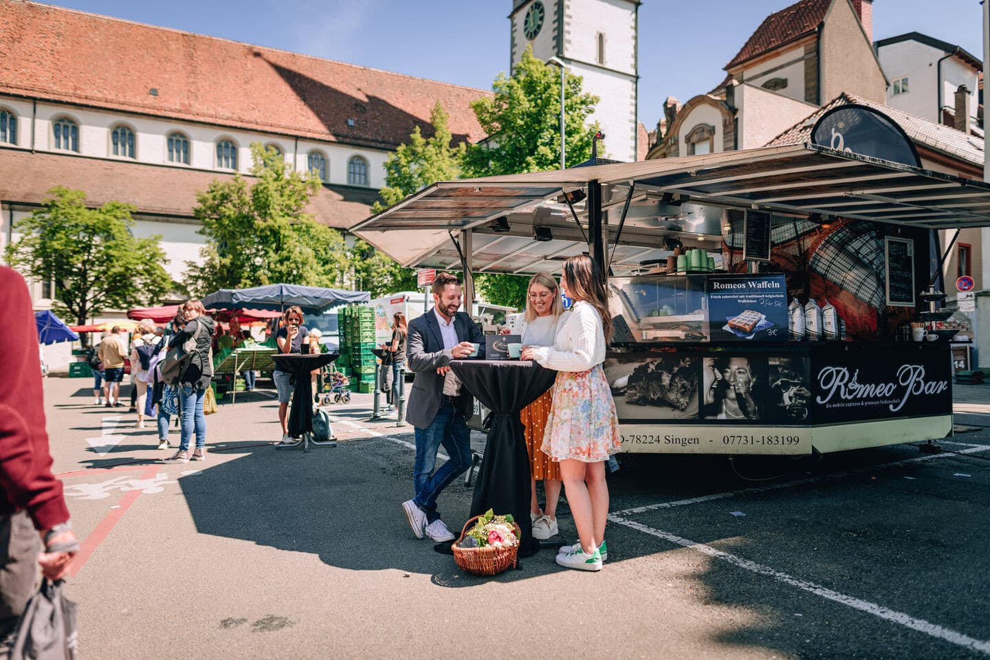 Eine Gruppe steht an einem sonnigen Tag beim Wochenmarkt auf dem Stephansplatz vor dem Stand der Romeo Bar und trinkt Kaffee.