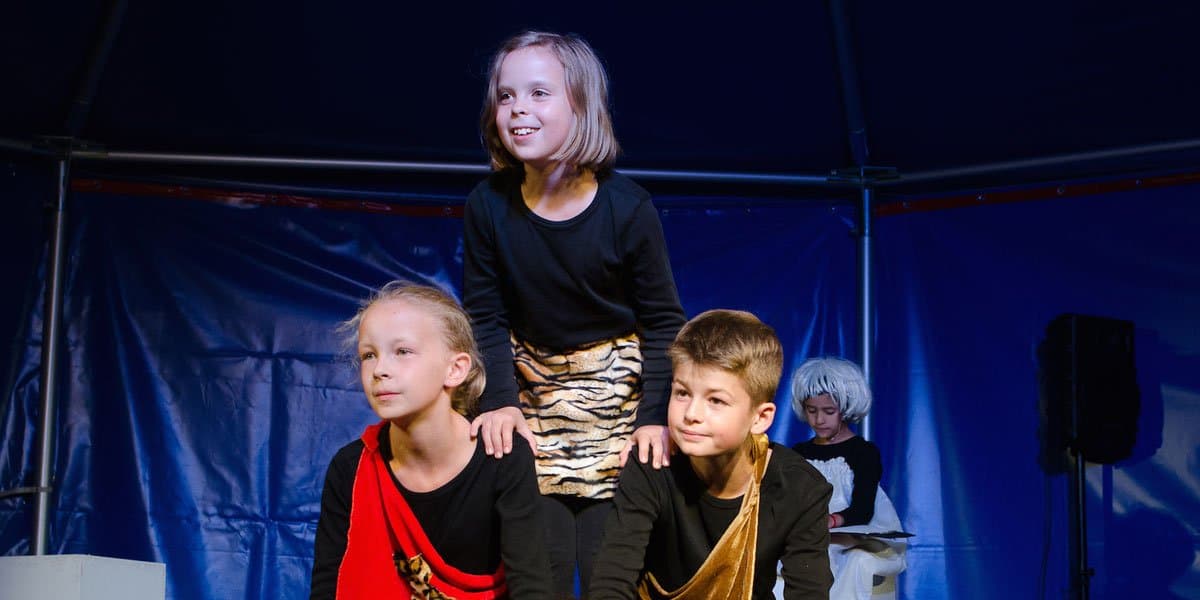 Drei Kinder in Kostümen auf einer Bühne, eines sitzt auf den Schultern der anderen beiden. Im Hintergrund ein weiteres Kind mit Perücke.