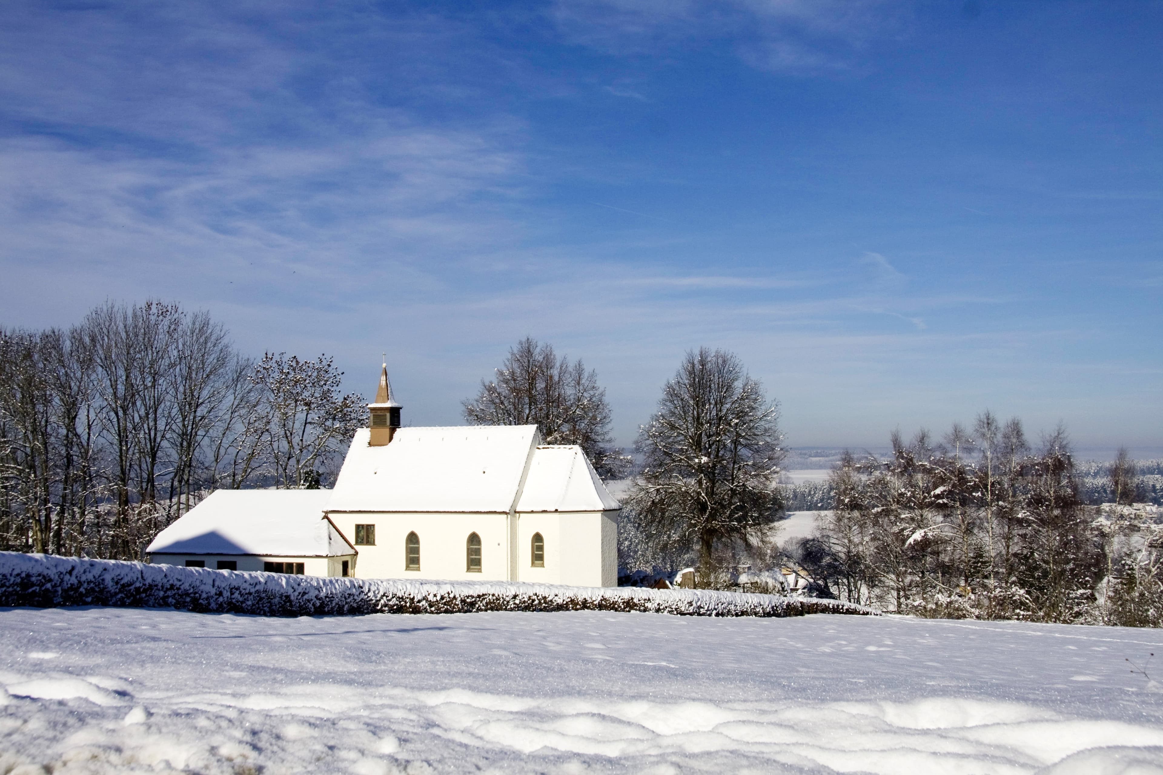 Auf dem Bild ist eine Kapelle mit verschneitem Dach und umgeben von einer winterlichen Landschaft zu sehen.