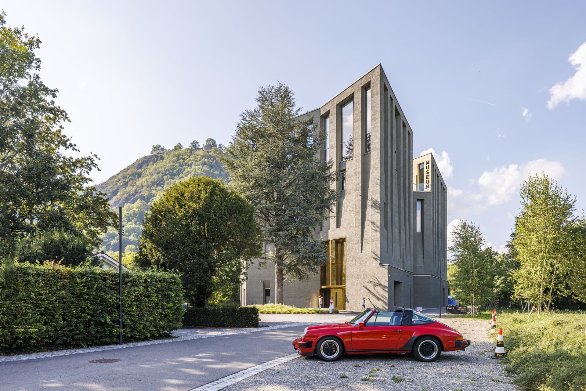 Modern museum complex with a red sports car at the front, surrounded by green spaces and mountains.