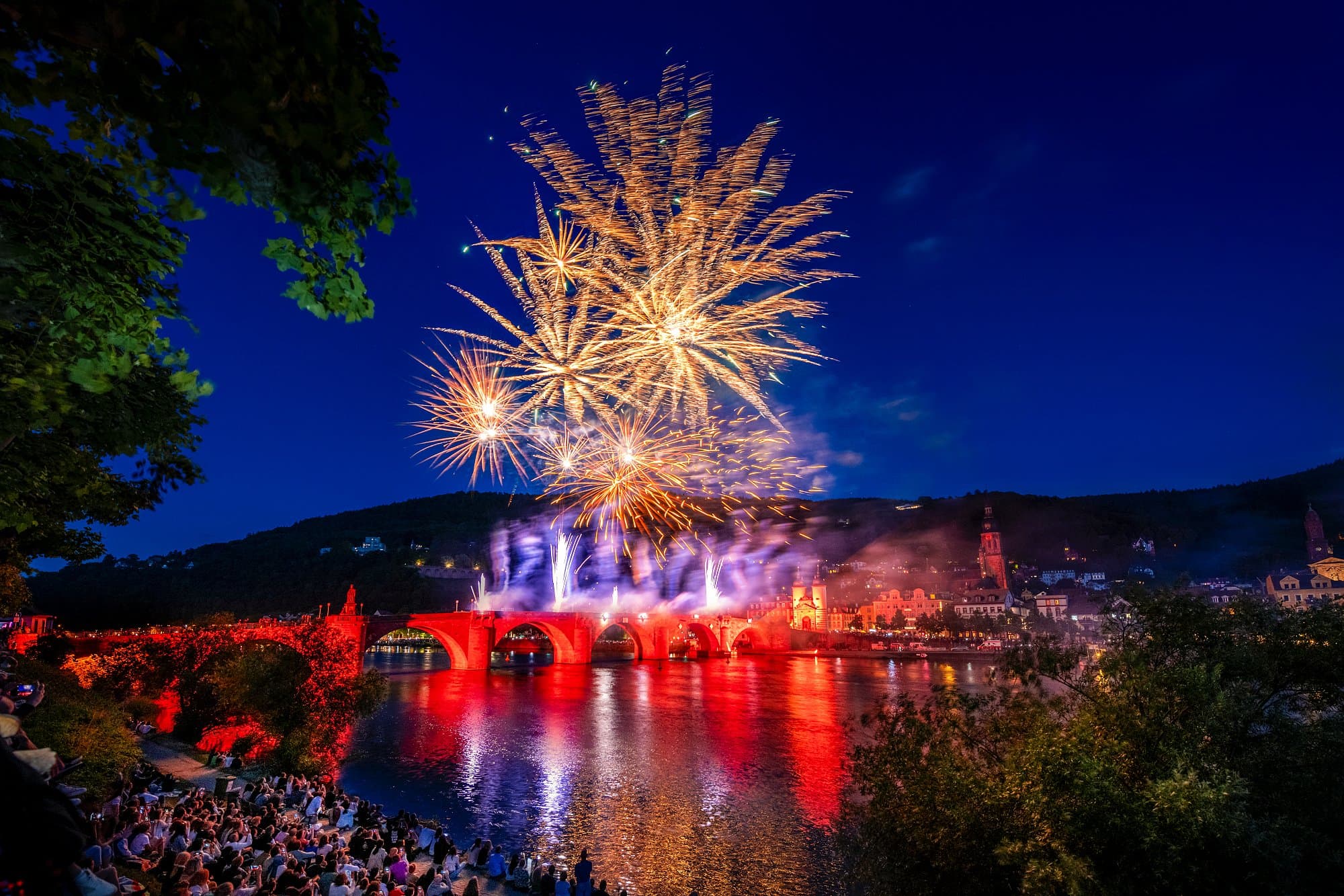Fireworks over the Old Bridge and Heidelberg Castle with spectators on the banks of the Neckar.