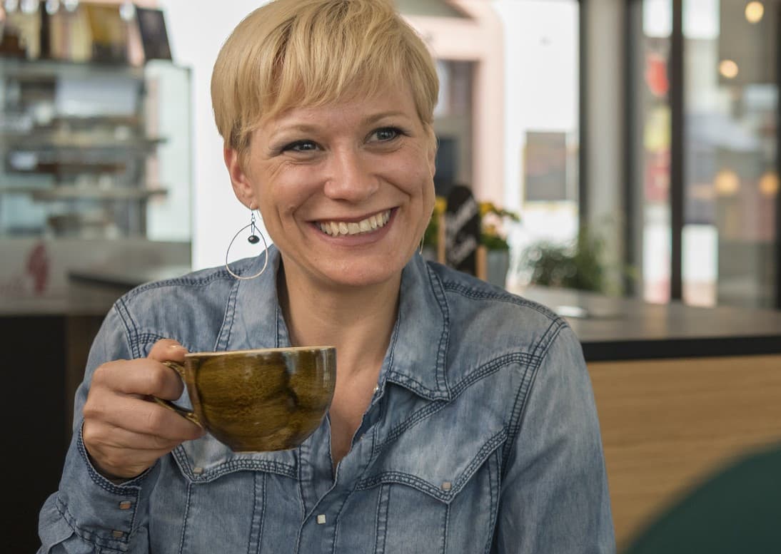 Auf dem Bild ist eine Frau mit kurzem, blondem Haar und hält eine Tasse in der Hand.