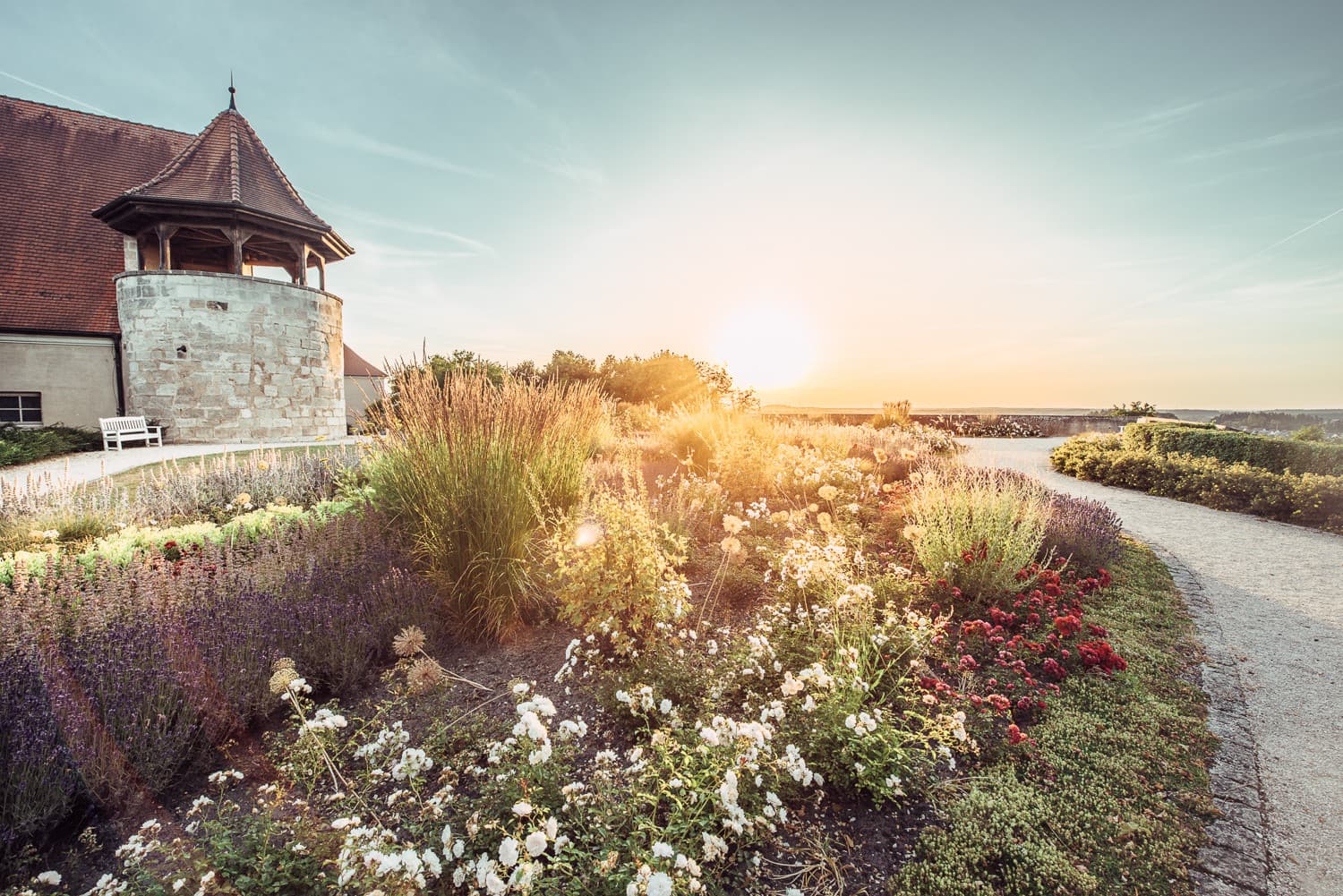 Sonnenuntergang über einem Garten mit blühenden Pflanzen neben einem historischen Turm und einem gepflasterten Weg.
