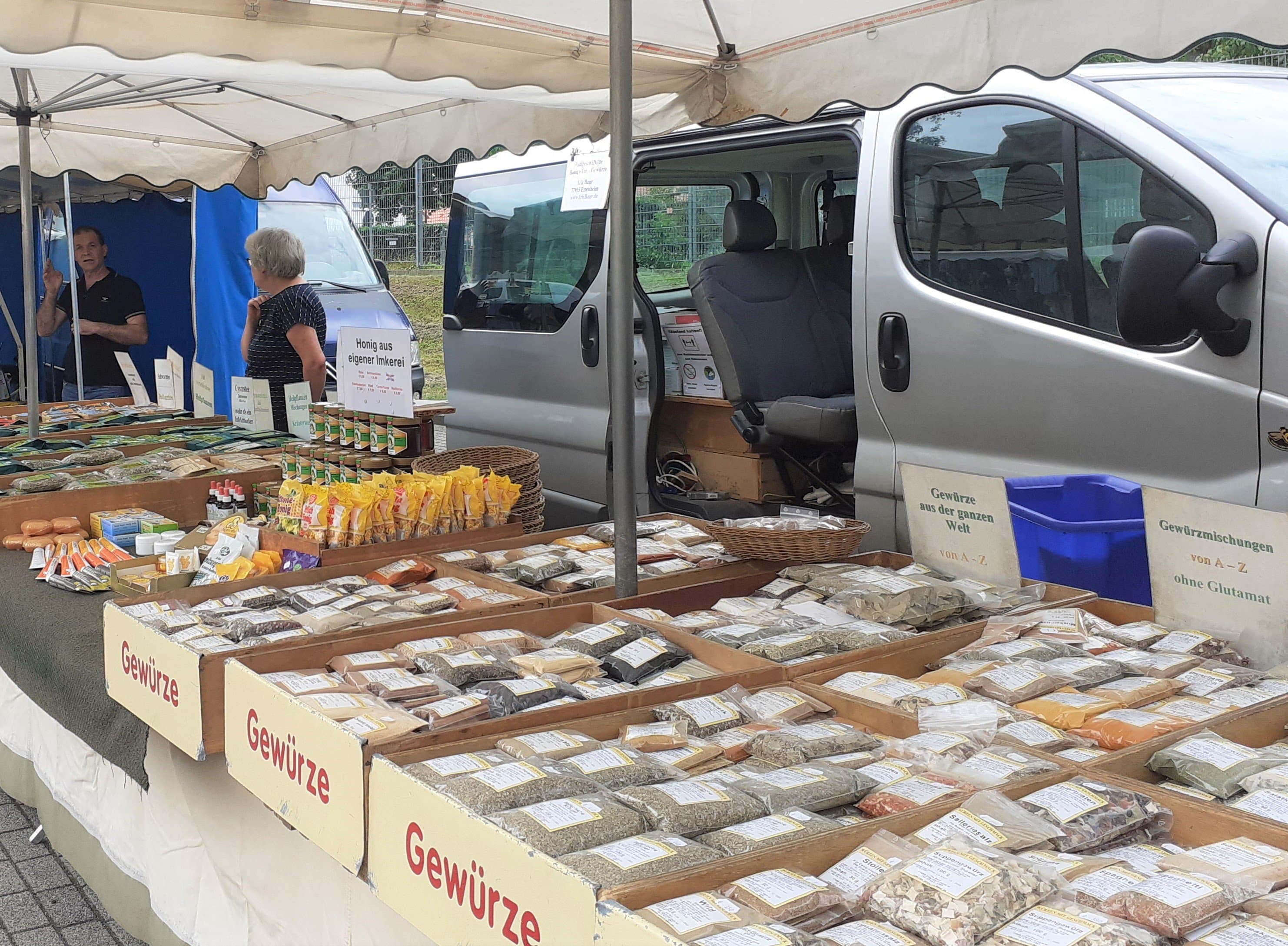 Man sieht einen Marktstand mit vielen Gewürzen. Im Hintergrund ein Transporter und 2 Verkäuferinnen.