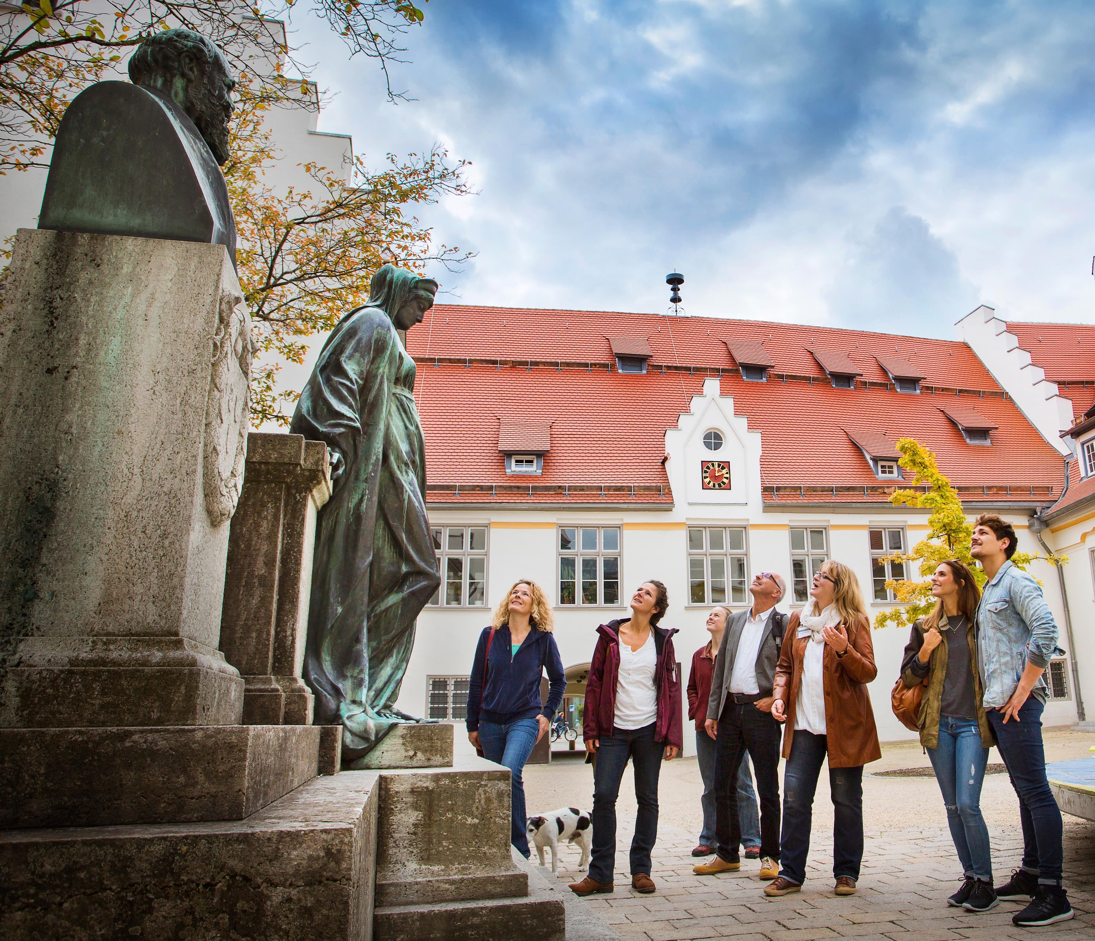 A group on a guided tour is looking at a bronze statue in front of the Biberach Hospital.