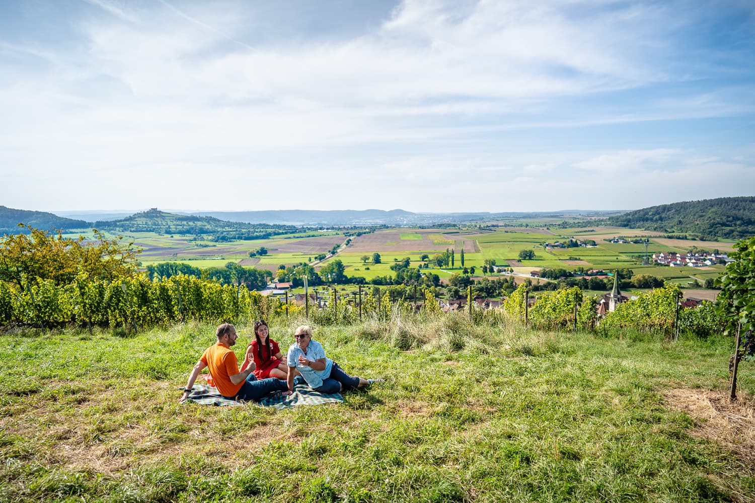 Drei Menschen auf einer Picknickdecke im Weinberg.