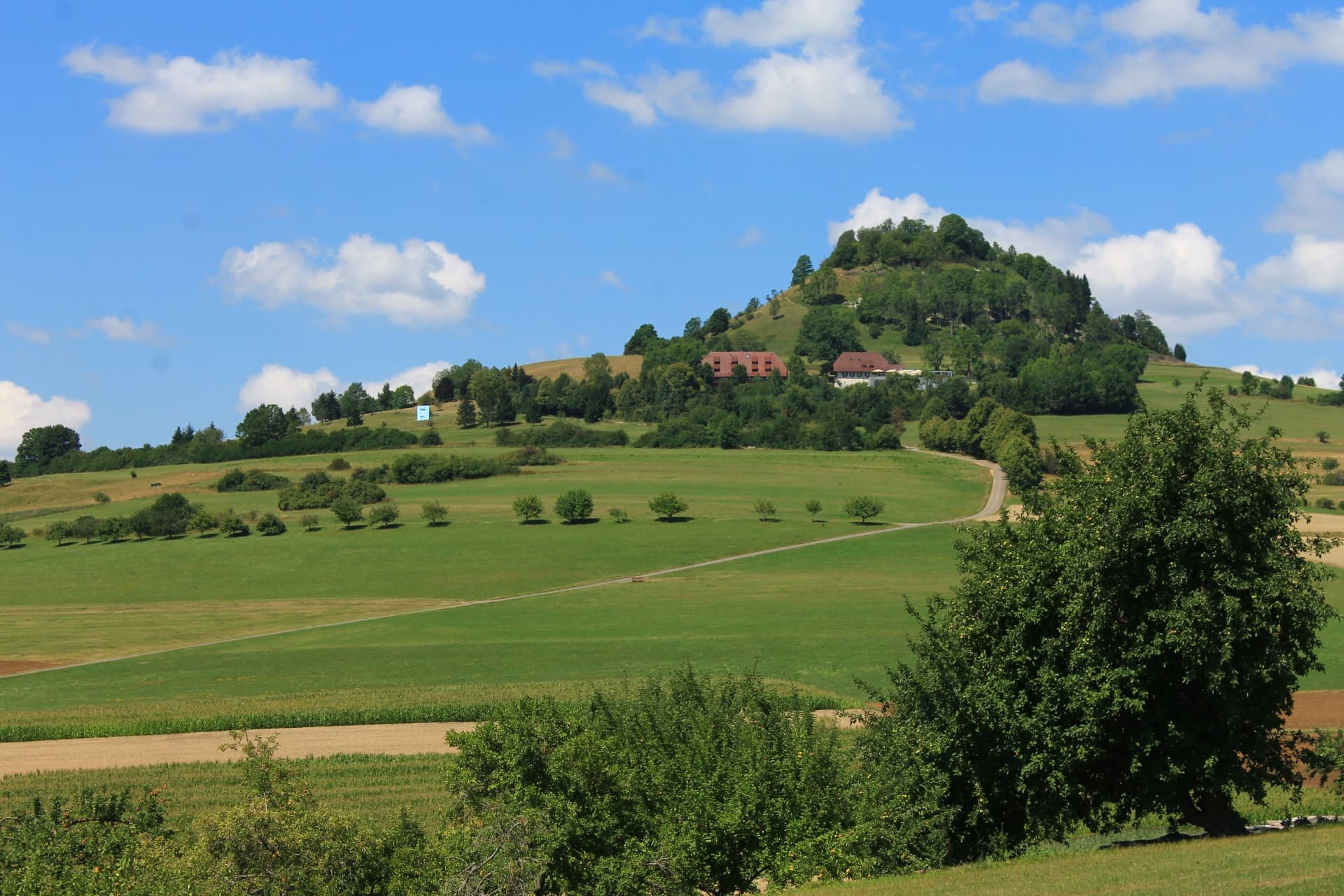 Blick auf kuppelförmigen, teilweise bewachsenen Hügel mit 2 Gebäuden vor blauem Himmel