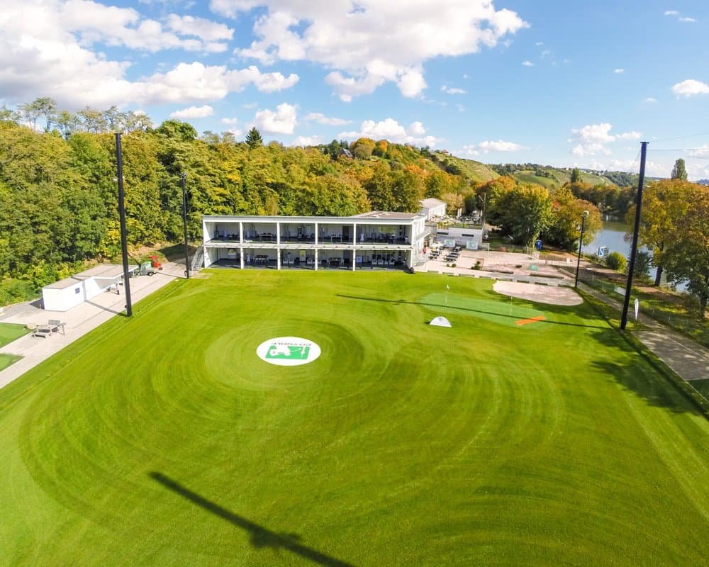 Luftaufnahme eines Golfplatzes mit Driving Range und Gebäude, umgeben von Bäumen.