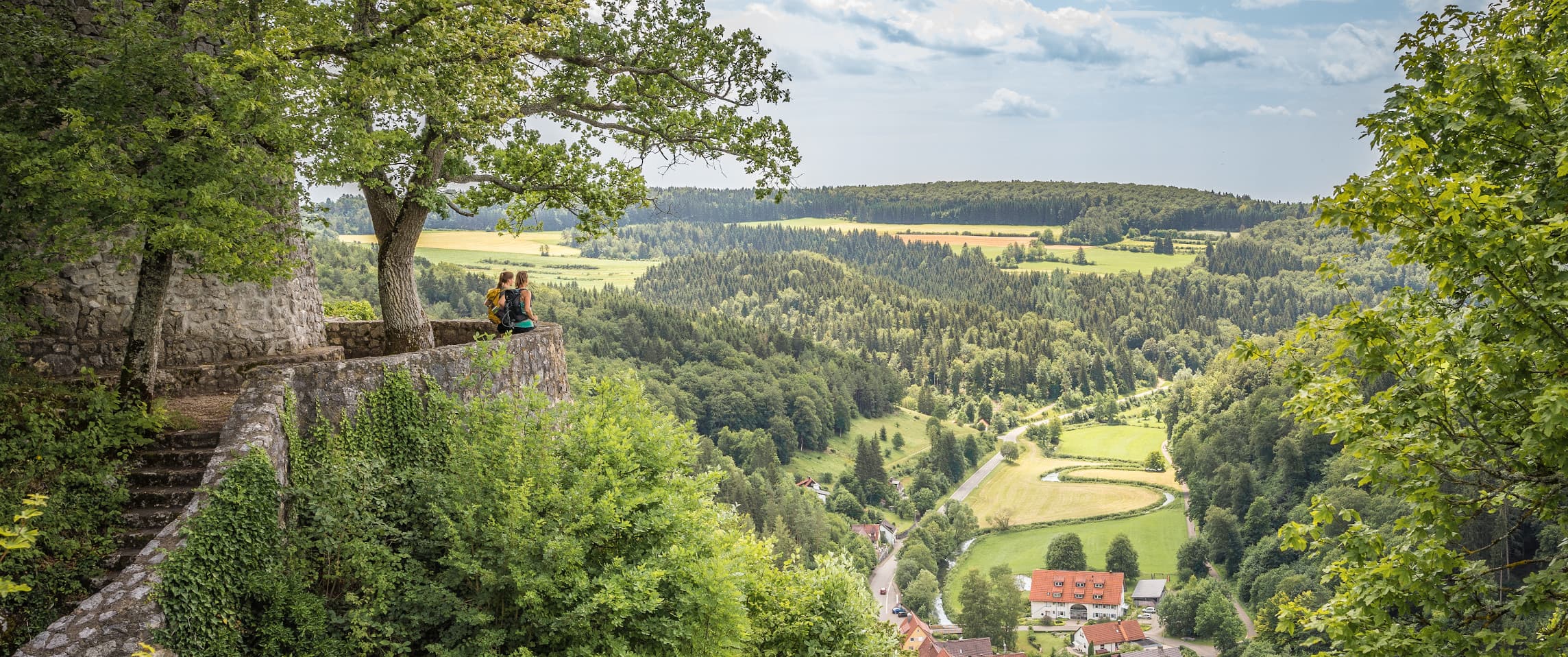 Die Burg Hohengundelfingen im Großen Lautertal in Münsingen im Biosphärengebiet Schwäbische Alb. Zwei Wanderer*innen stehen auf einer Plattform an einer Burg. Sie blicken ins Tal durch das sich ein Fluss schlängelt und ein kleines Dorf liegt.