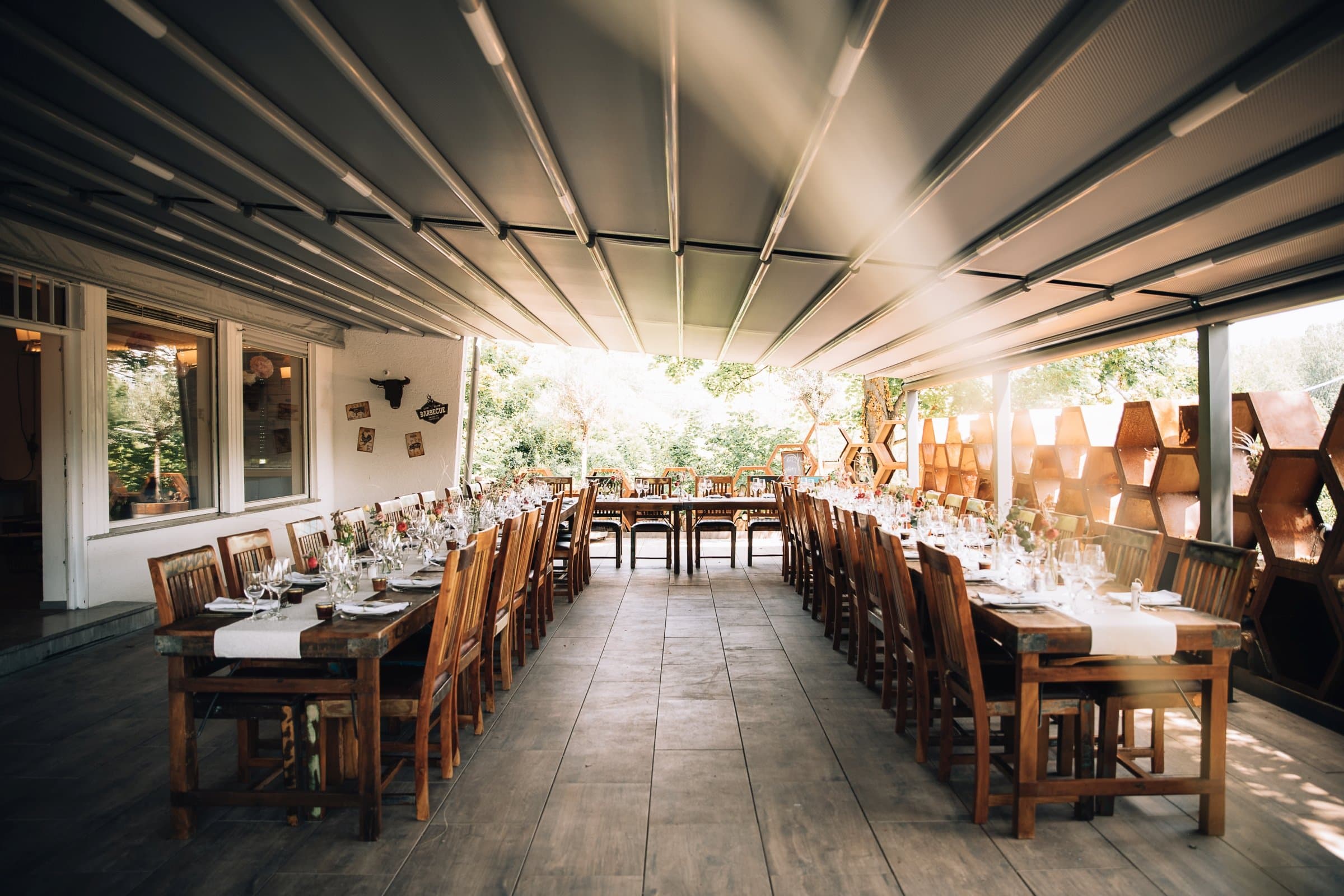 Überdachte Terrasse mit langen Holztischen und Stühlen, gedeckt für ein Essen, umgeben von Bäumen und dekorativen Wänden.
