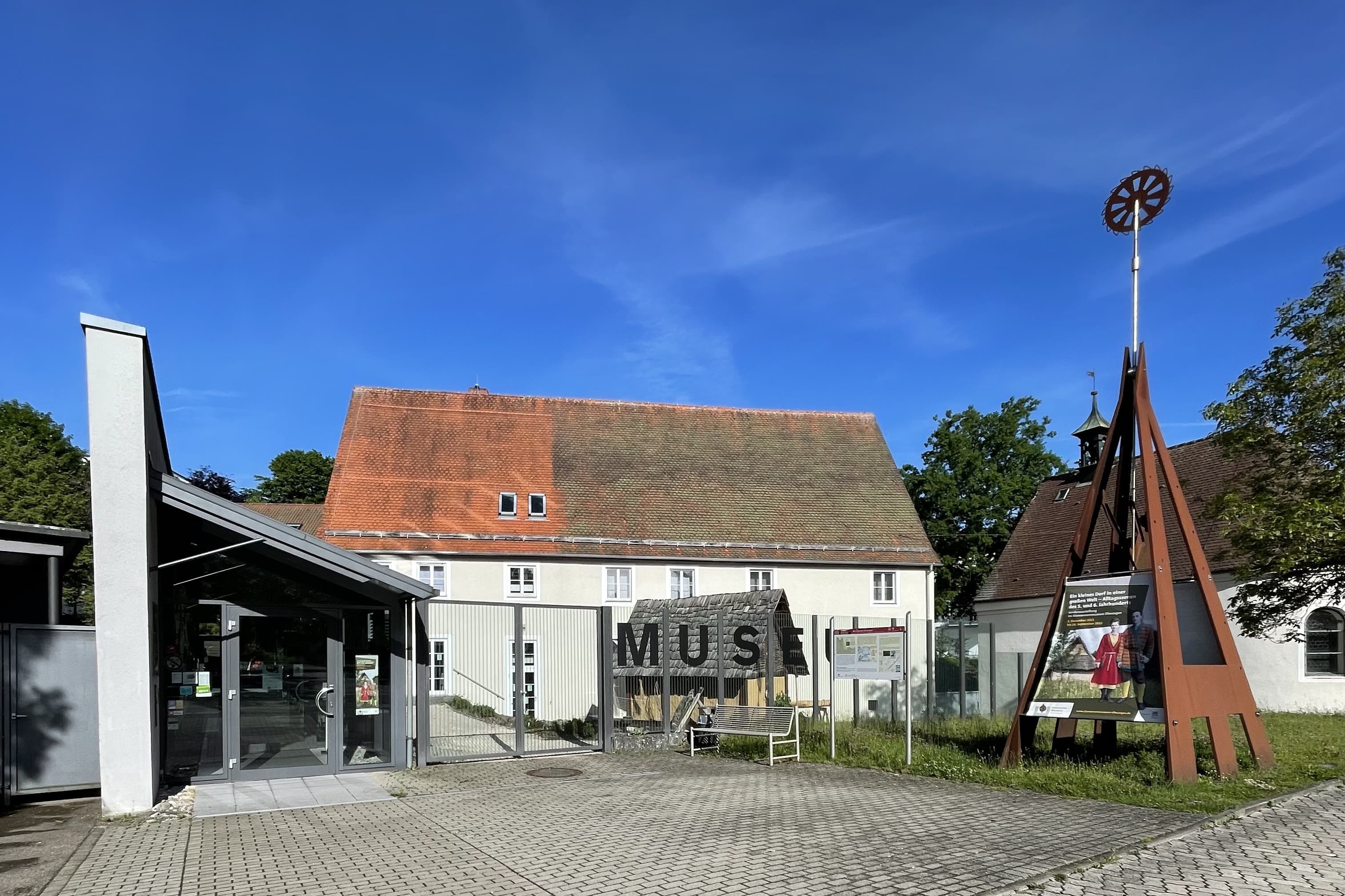 Blick auf Gebäude. Davor ein Zaun mit Aufschrift Museum. Daneben Dreieckige Skulptur aus Metall  mit kleinem Rad.