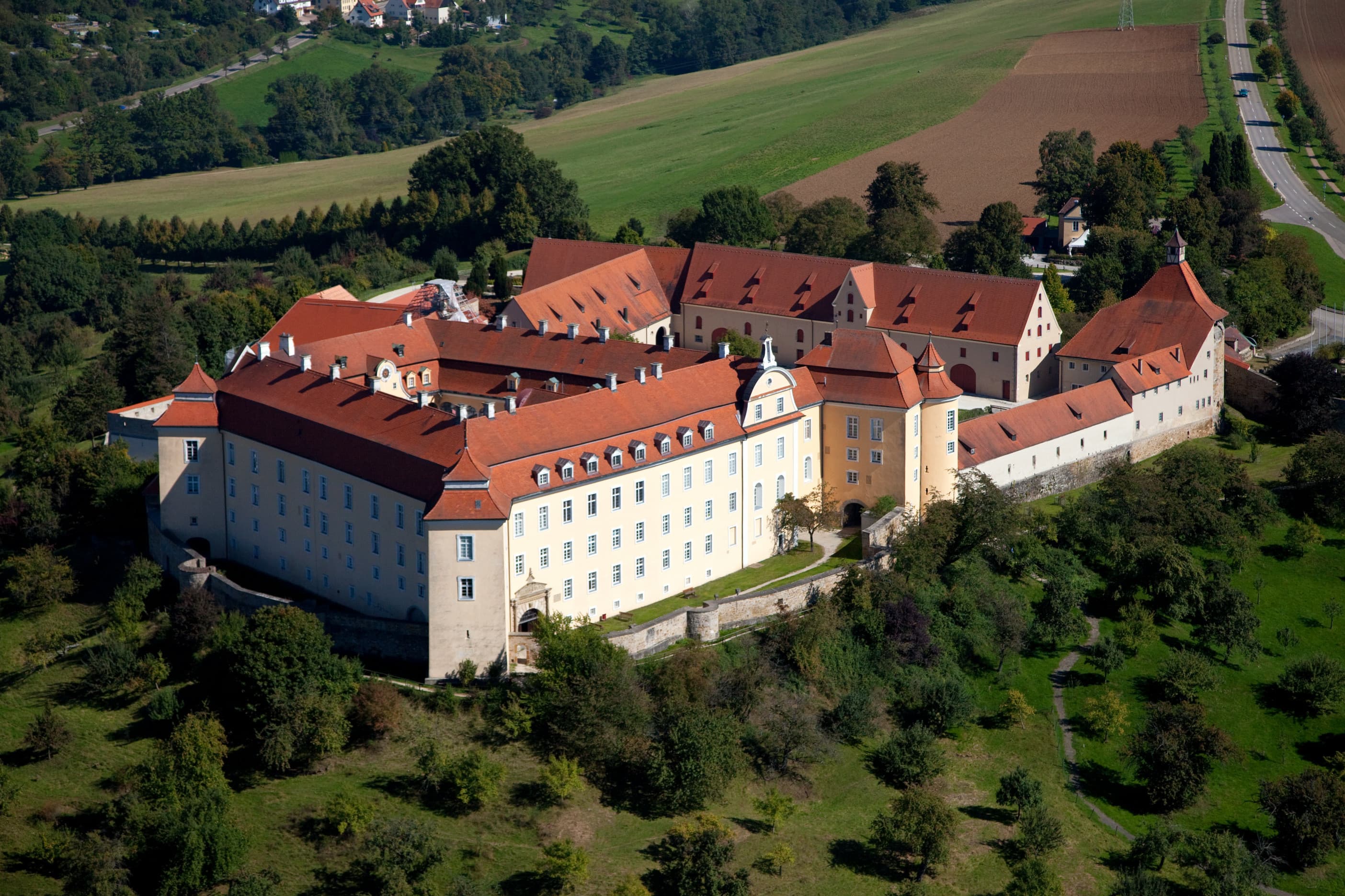 Luftbild großes, historisches Schloss mit roten Ziegeldächern, umgeben von Bäumen und Feldern.