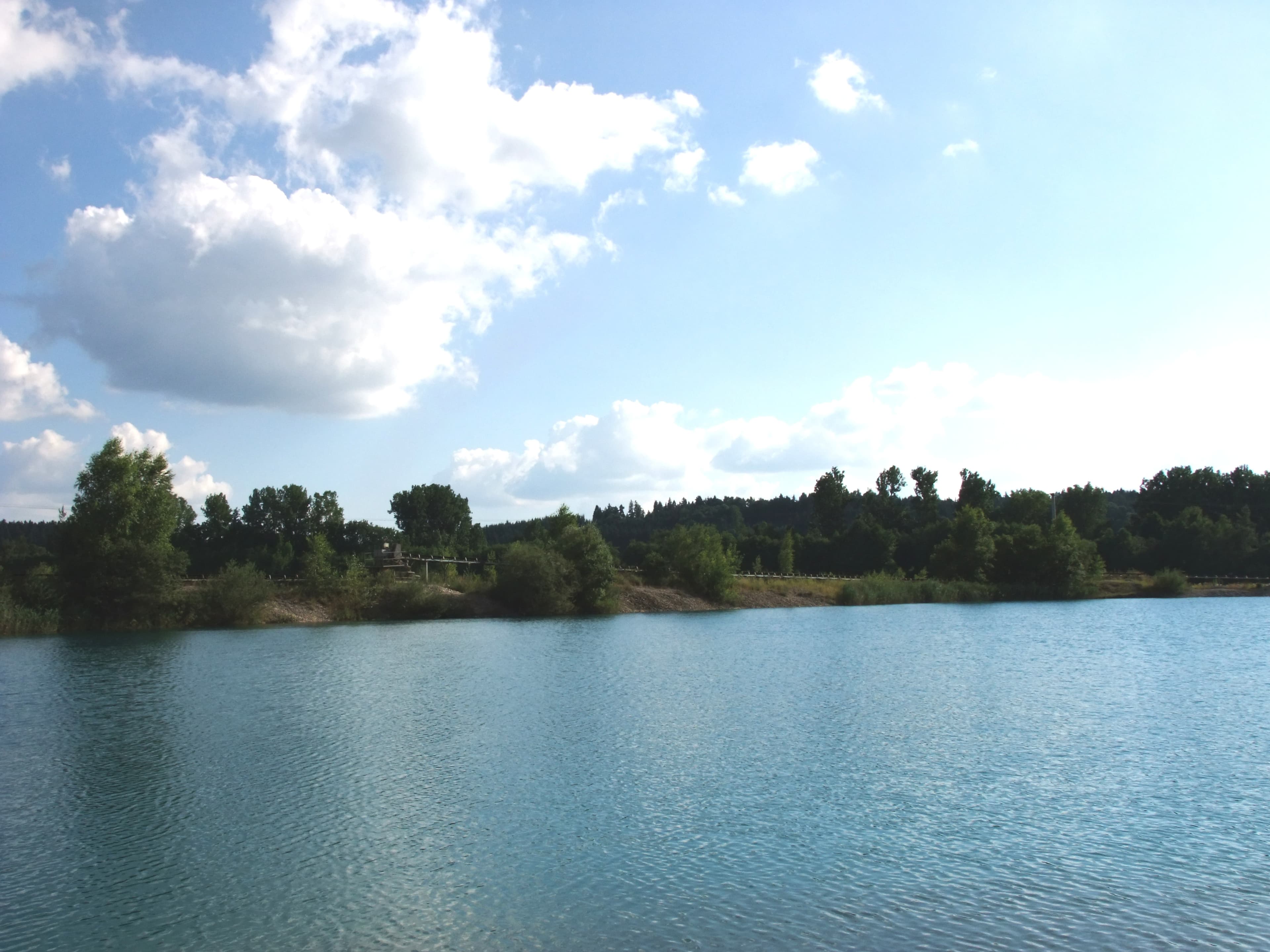 Blick über den See zum grün mit Bäumen bewachsenen Ufer unter blauem Himmel mit Wolken
