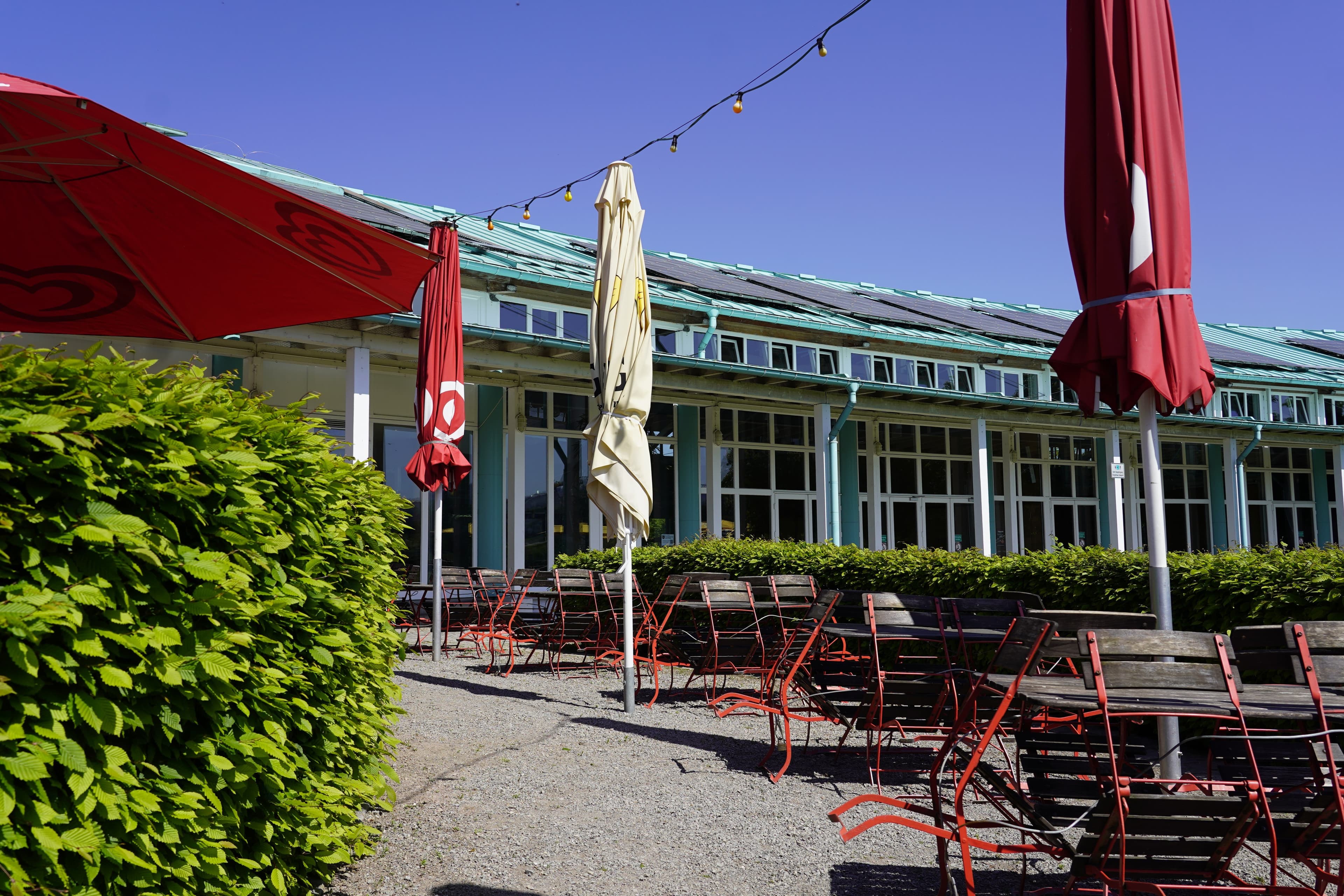 Außenbereich eines Restaurants mit roten Sonnenschirmen und leeren Stühlen vor einem Gebäude mit großen Fenstern.