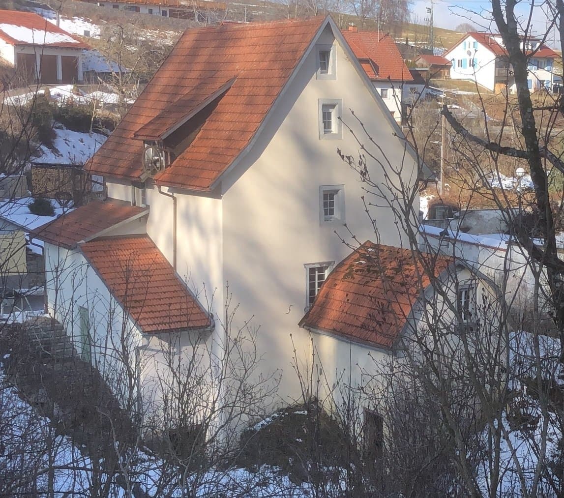 A white house with a red roof and several dormers in a snowy landscape.