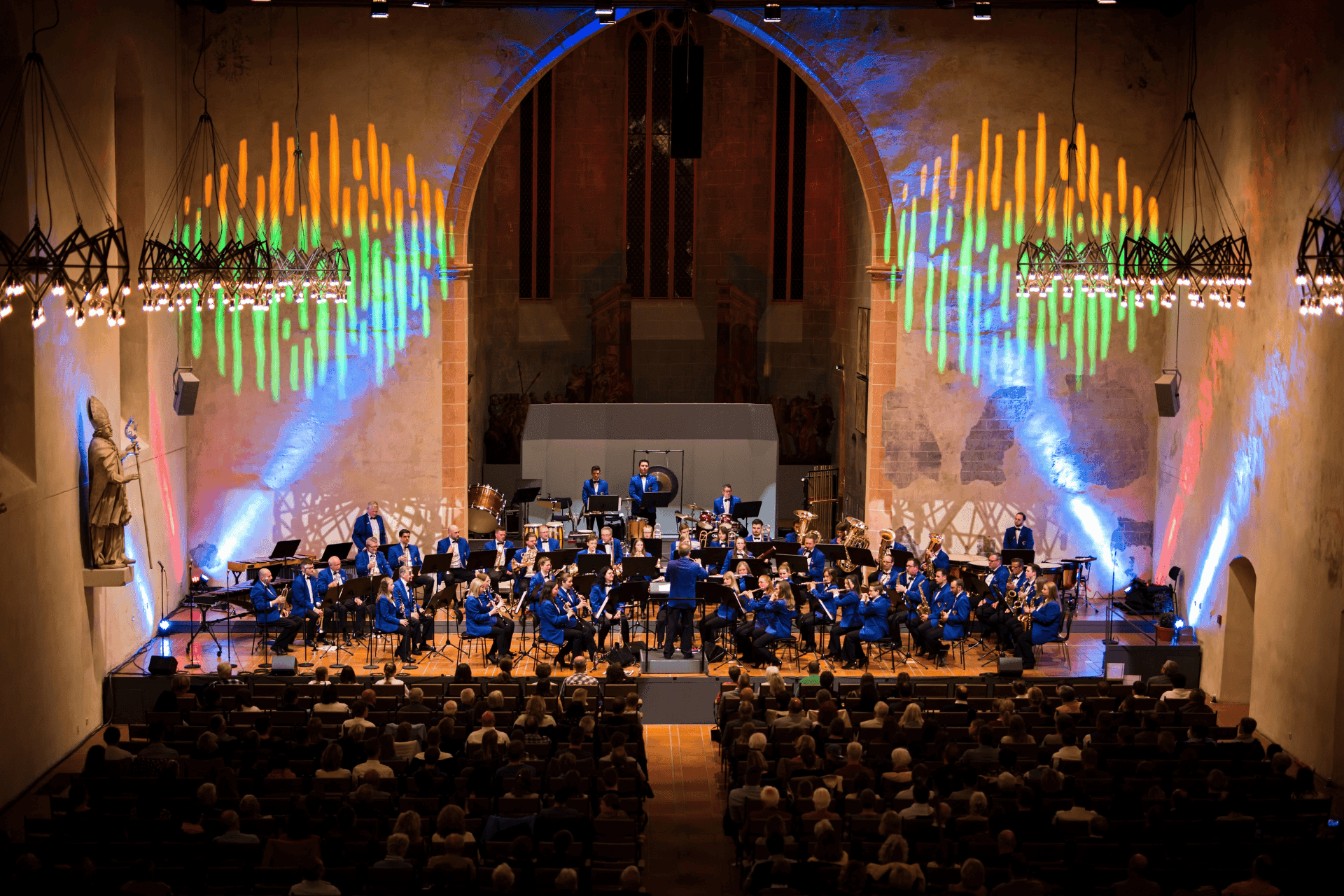 Orchesterauftritt in einer großen Kirche: Musiker mit Streichern, Bläsern und Schlagwerk auf der Bühne vor Publikum, bunte Lichtprojektionen an den Kirchenwänden.