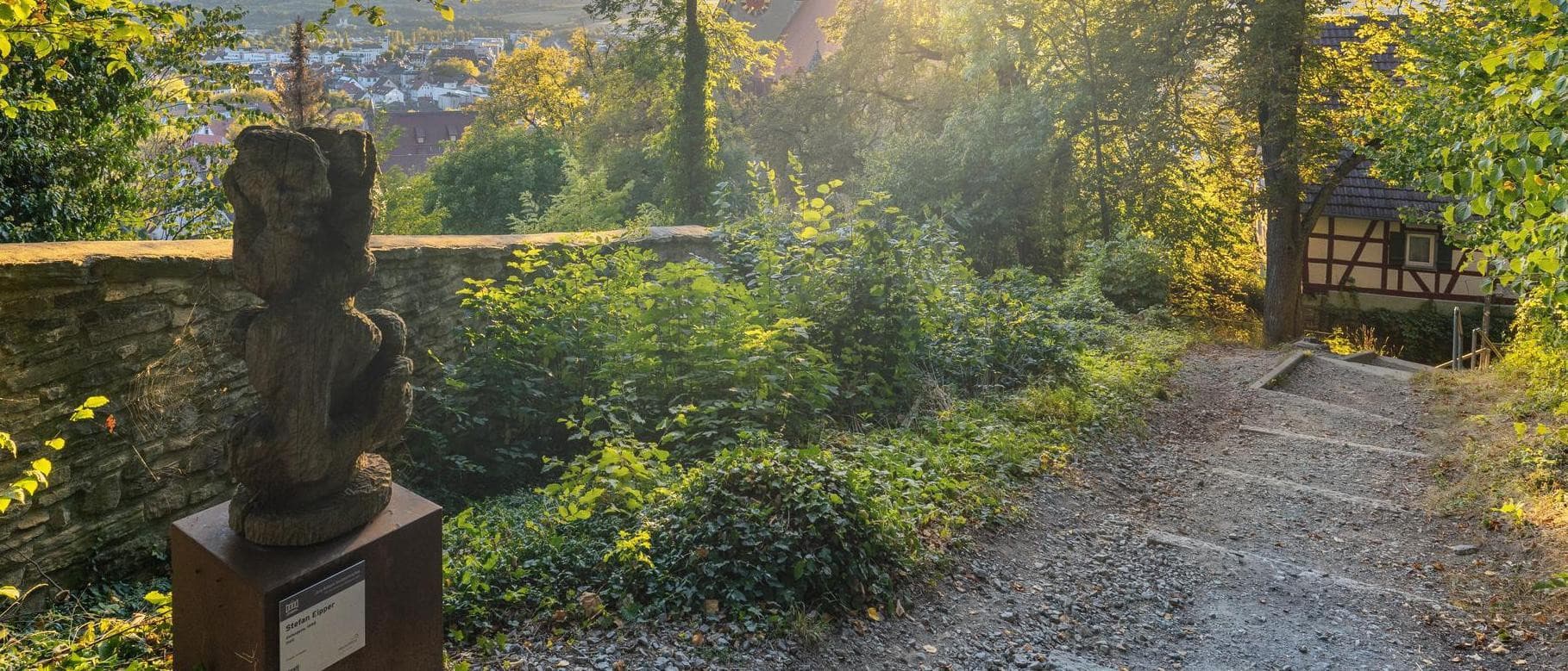 Skulptur auf einem Pfad in Herrenberg, umgeben von Bäumen und einem Fachwerkhaus im Hintergrund.