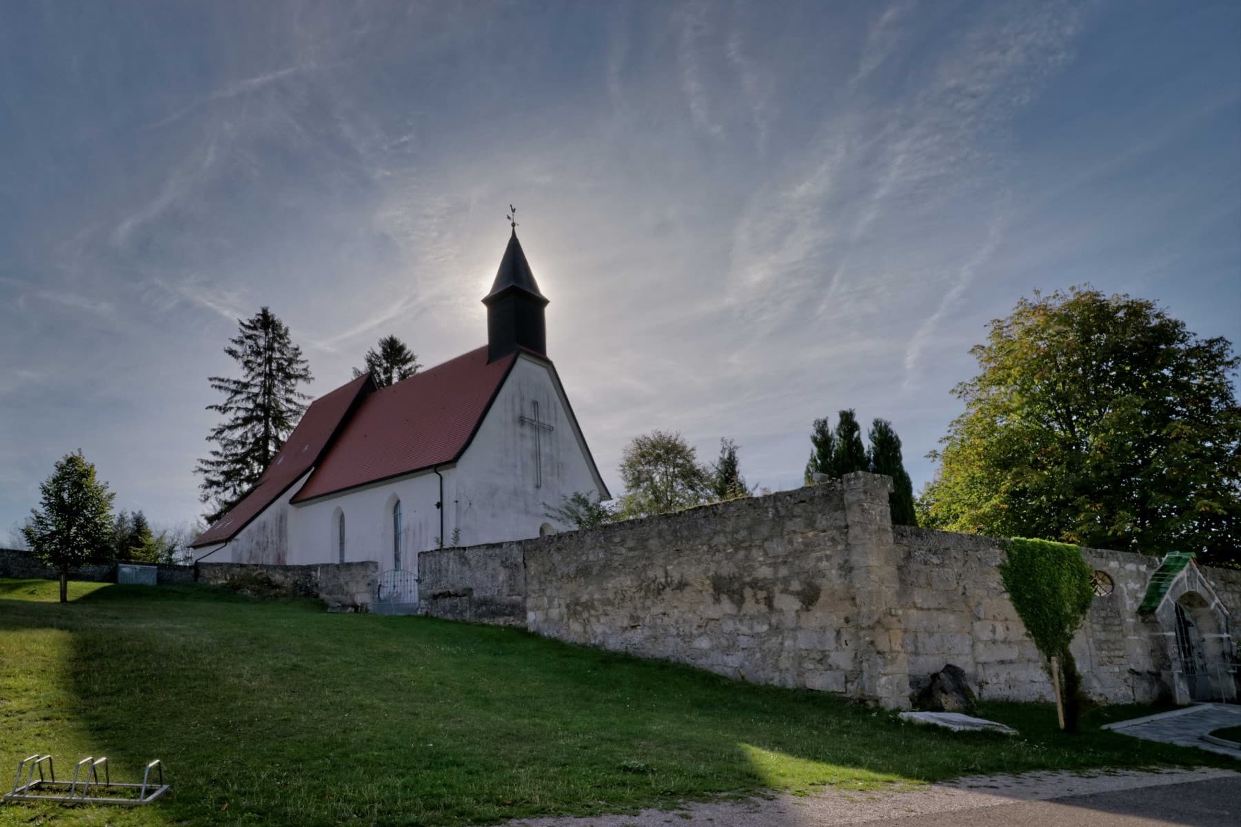 Gruorn im ehemaligen Truppenübungsplatz in Münsingen im Biosphärengebiet Schwäbische Alb. Eine Kirche, hinter deren Kirchturm die Sonne strahlt. Die Kirche hat ein großes Kreuz auf der Fassade. Davor ist eine Steinmauer.