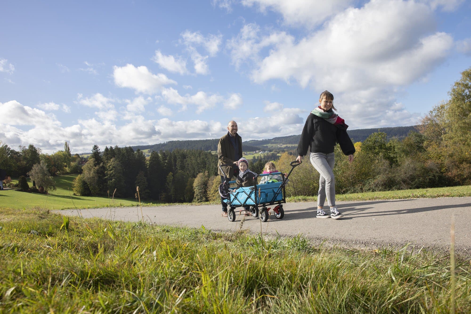 The picture shows a family on the AugenBlick-Runde Sulgen. The two children are sitting in a blue handcart and in the background you can see the distant view of the Black Forest landscape.