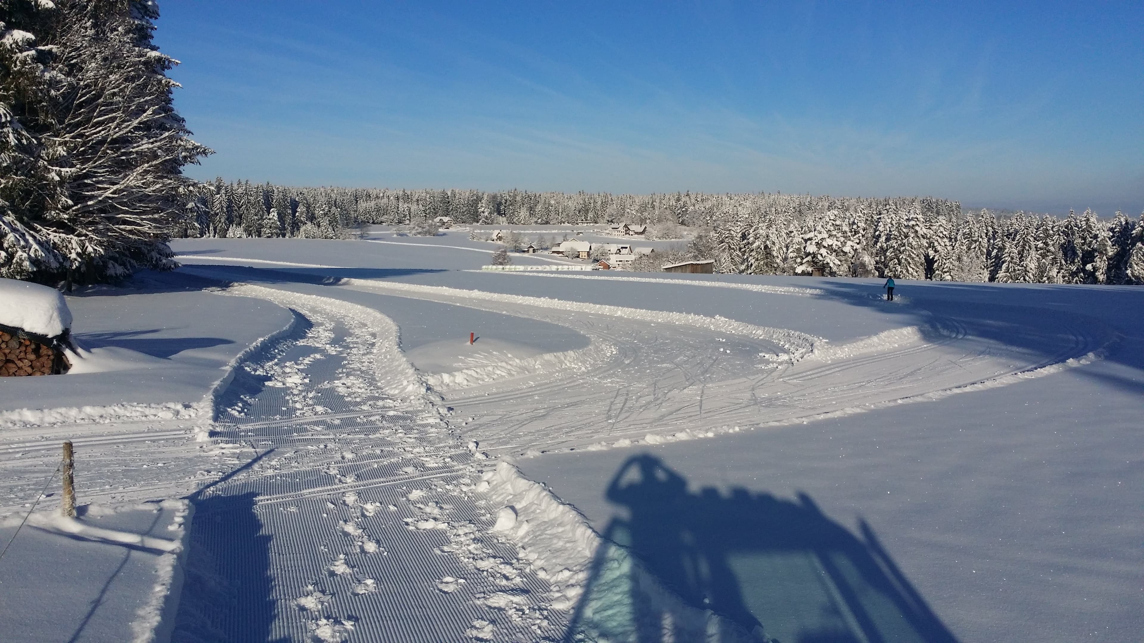 Das Bild zeigt eine schneebedeckte Landschaft mit einer Langlaufpiste und einem gespurten Winterwanderweg auf der Ramsteiner Höhe. Im Hintergrund sieht man eine verschneite Schwarzwaldlandschaft.
