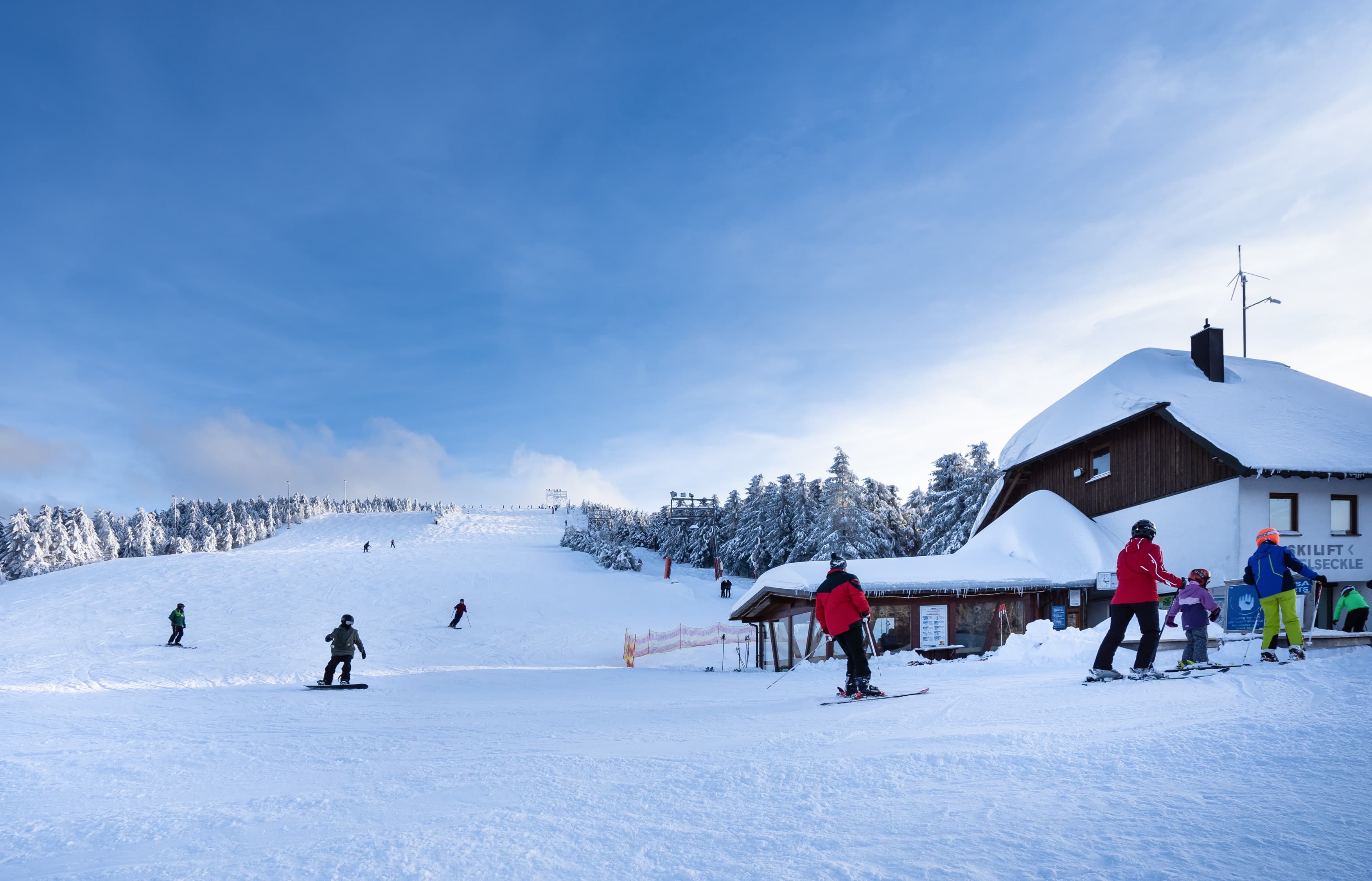 Blick auf den Skihang Seibelseckle mit Menschen auf Skier und rechts das Skiliftgebäude mit Skibar. Strahlende Sonne und blauer Himmel.
