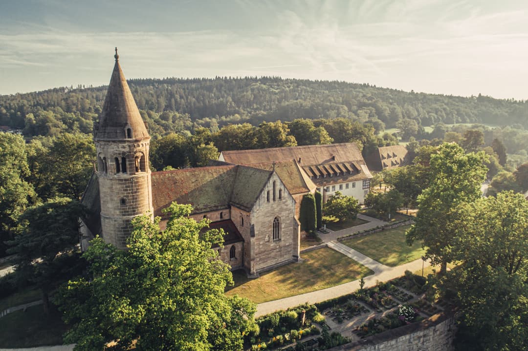Aerial view of a historic monastery with a round tower and surrounding forest in soft afternoon light.