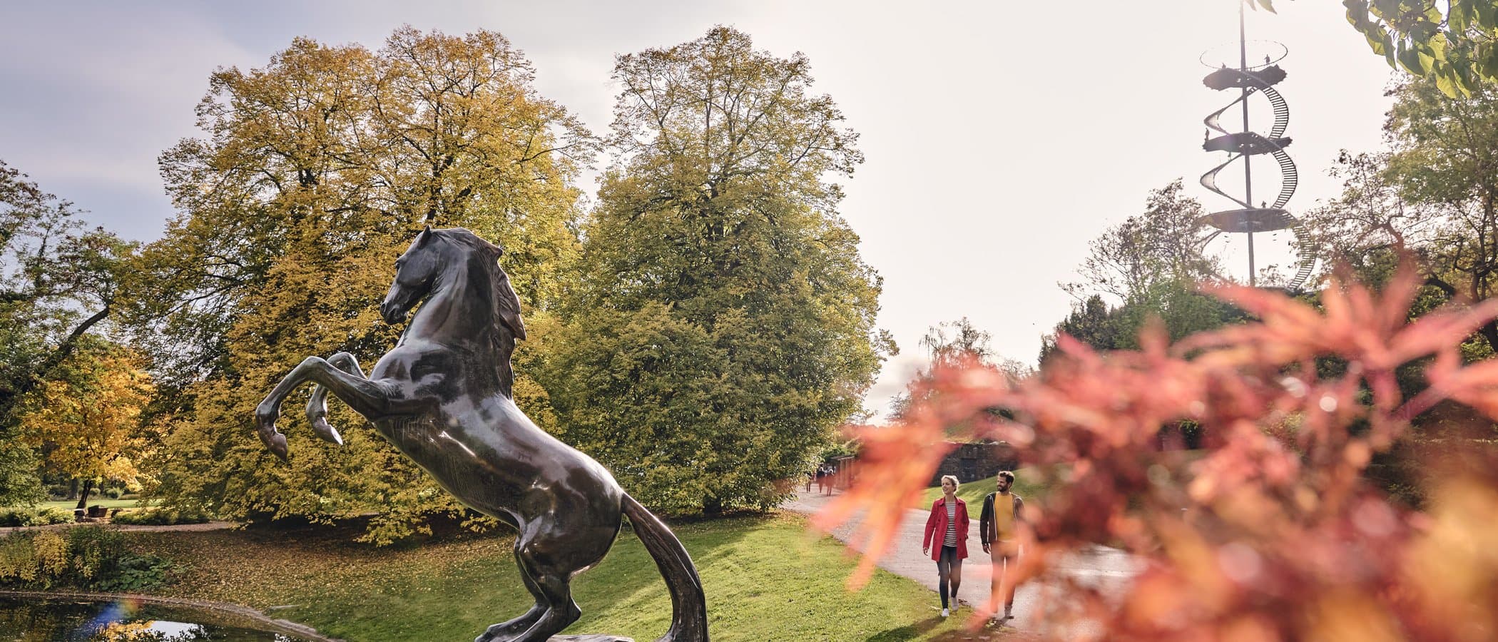Bronzestatue eines Pferdes im Höhenpark Killesberg, Stuttgart, mit Herbstbäumen und einem spiralförmigen Aussichtsturm im Hintergrund.