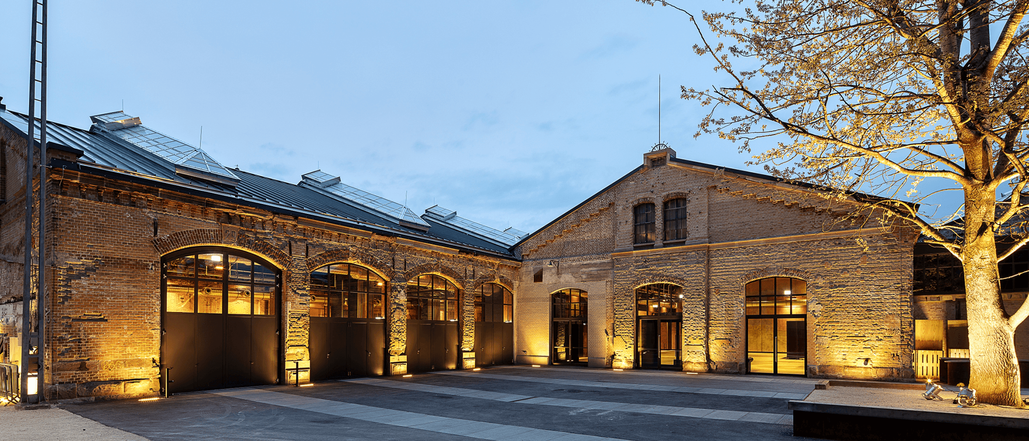 Außenansicht der beleuchteten Wagenhallen mit Backsteinfassade und Glasdach bei Dämmerung.