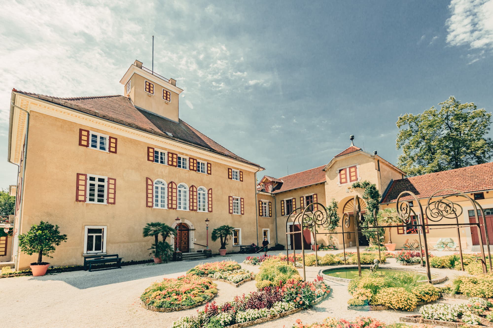 Gelbes Schloss mit roten Fensterläden und einem Garten mit Blumenbeeten im Vordergrund unter blauem Himmel.