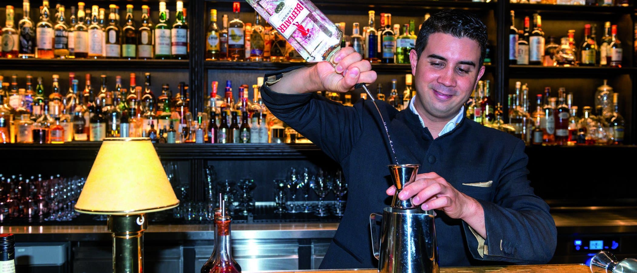 Ein Barkeeper gießt Gin in einen Jigger vor einer Bar mit vielen Flaschen.