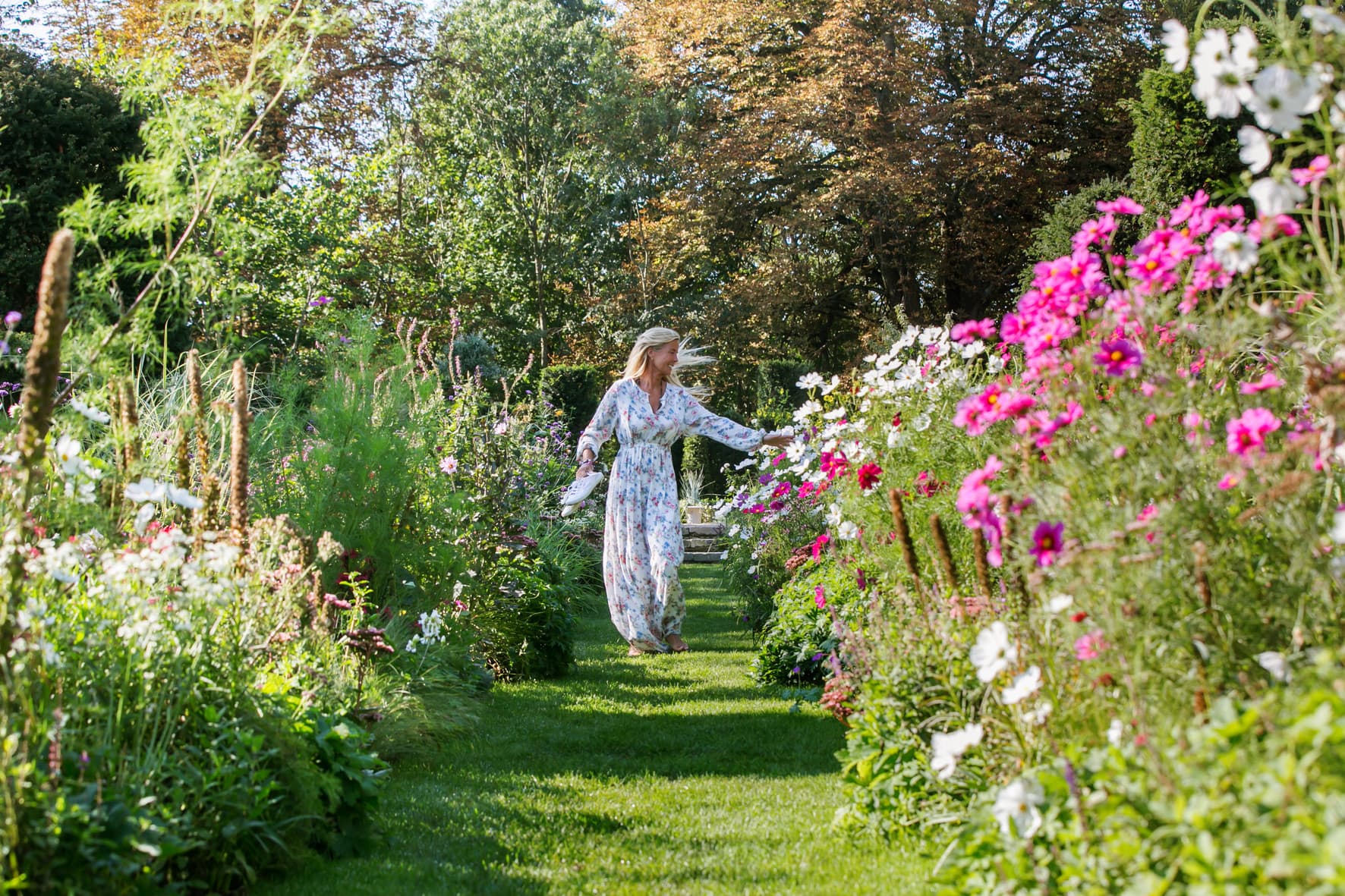 Frau in langem, hellblauem Kleid geht durch blühenden Garten eines Schlosses mit bunten Blumen.